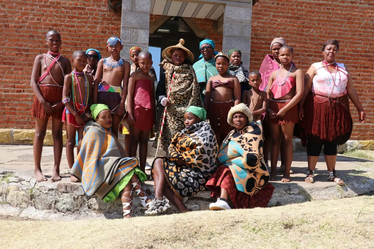 Community members and young performers in traditional Basotho dress gather outside a sandstone hall.